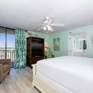 The master suite features a cozy armchair, a TV, and tropical decor, with a lovely view from the balcony