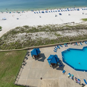 The view showcases a vibrant pool area with blue loungers and cabanas, leading to a beautiful beach with white sand