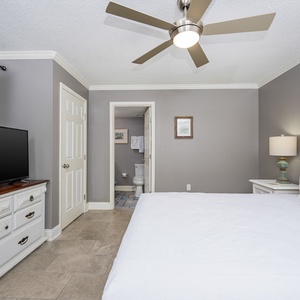 Bright guest bedroom featuring a queen bed, large TV, and a view into the bathroom area with elegant decor and tile flooring