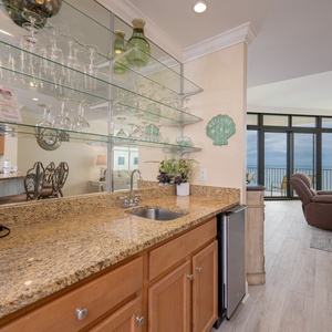 A stylish wet bar area features granite countertops, a sleek ice-maker, and a welcoming view of the expansive coastline