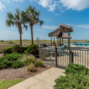 Lush greenery surrounds the pool area, featuring a shaded gazebo and lounge chairs with a view of the beach in the distance