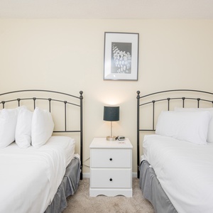 Guest bedroom features two full beds with white linens, nightstand with a lamp, and soft beige walls for a serene atmosphere