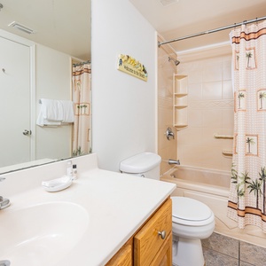 The bathroom features a light-colored vanity, a tub/shower combo, and a palm-patterned shower curtain for a tropical touch