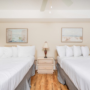 This guest bedroom features two neatly made full beds, a bedside table with a lamp, and coastal-themed artwork on the walls