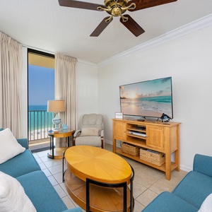 Living area features turquoise sofas, a dining table, and a large TV, with views of the beach from the balcony