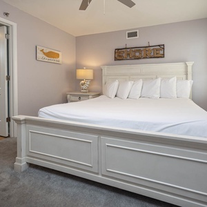 A serene guest bedroom features a plush king bed with multiple pillows, soft gray carpet, and beach decor on the walls