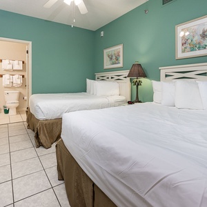 Bright teal walls complement two queen beds with white linens, a palm-themed lamp, and an en-suite bathroom with fresh towels