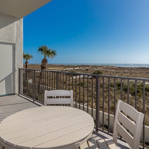 Balcony with a round table and chairs overlooks lush greenery and the tranquil gulf, perfect for enjoying the view