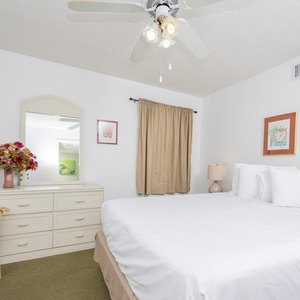 A bright guest bedroom features a queen bed, decorative artwork, wicker chair, and a dresser with a mirror and fresh flowers