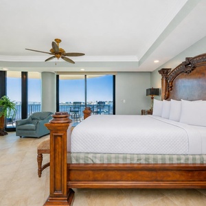 The grand master bedroom features a king wooden bed, a baby grand piano, and view of the bay through floor-to-ceiling windows