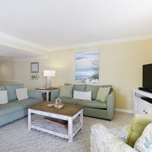 Cozy living area features soft green sofas with queen sleeper, rustic coffee table, and large TV, surrounded by beach decor
