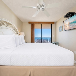 Bright master bedroom with a king bed, a mounted TV, and sliding glass doors leading to a private balcony with beach views
