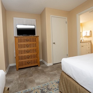 Guest bedroom featuring two double beds, a woven dresser with a TV, and an adjoining bathroom visible through the door