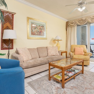 The living area features a cozy seating arrangement, vibrant decor, and a stunning beach view through large sliding doors