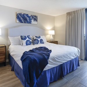 Inviting master suite featuring a king bed with blue accents, a side table lamp, and a balcony offering a view of the beach