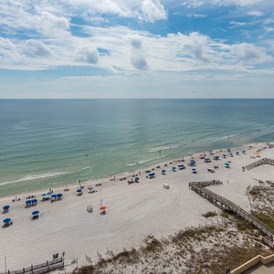 The image captures a serene beach front with white sand, colorful umbrellas, and gentle waves under a bright blue sky