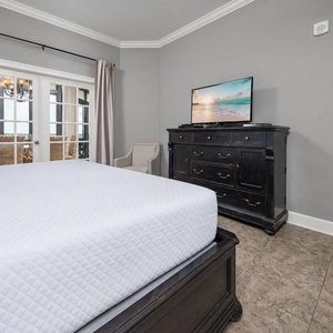 The master features a king bed, sleek black dresser, a TV, and elegant curtains framing the view of the beachfront