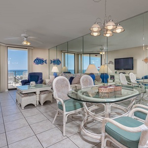 Bright living area features a glass dining table, comfortable seating, and a panoramic view of the beach from large windows