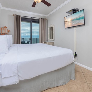 Master suite bedroom featuring a comfortable king bed, a mounted TV, and access to a balcony with a view of the beach