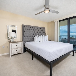 Cozy master bedroom featuring a plush king with gray tufted headboard, light bedding, and a balcony with a view of the beach