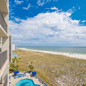 Balcony view showcases the pool area and expansive beach with white sand, framed by vibrant blue skies and gentle waves