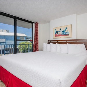 The bedroom features a king bed with crisp linens, vibrant red accents, and a view of the balcony through expansive windows