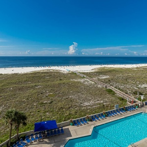 Expansive view of the beach with white sand, a pool area featuring blue loungers, and a clear blue sky above