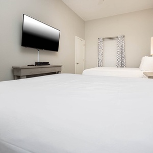 Bright guest bedroom featuring two double beds, a mounted TV, and soft linens, creating a welcoming and restful atmosphere