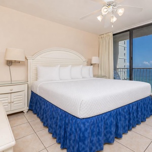 The master bedroom features a plush king bed with a blue skirt and a stunning beach view through large sliding glass doors