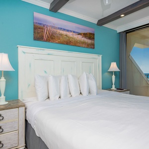 The master bedroom features a serene turquoise wall, plush bedding, and a stunning beachfront view through large windows