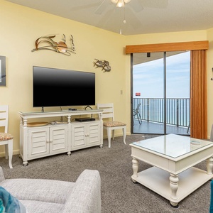 Living area features a large TV, comfortable seating, and sliding glass doors opening to a balcony with beach front views