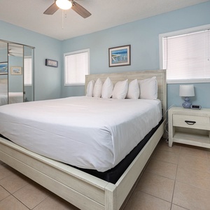 The master bedroom features a comfortable bed with plush pillows, light blue walls, and ample natural light from the windows