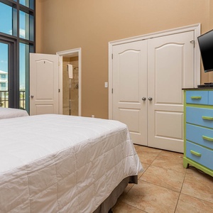 This bright bedroom features two full beds, a colorful dresser, and a TV, with large windows offering a view of the outdoors