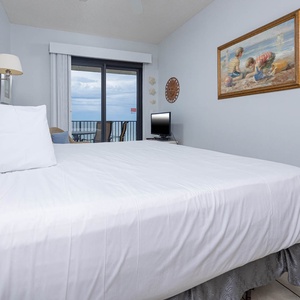Master bedroom features a large king bed, a TV, and a balcony offering a glimpse of the gulf view beyond