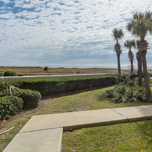 Lush greenery frames a pathway leading to a pool area with blue umbrellas, offering a serene view of the distant beach