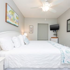 Cozy guest bedroom featuring a king bed with decorative pillows, a wall-mounted TV, and a colorful beach-themed painting