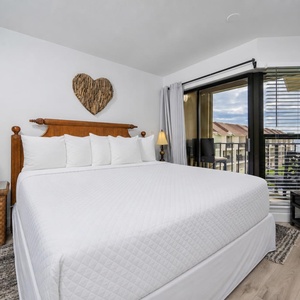 The master bedroom features a king bed with plush pillows, a charming lamp, and a view of the balcony area through large windows