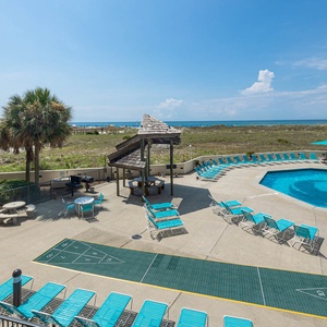 Relax by the pool area featuring turquoise lounge chairs, a shuffleboard game area, and a view of the beach in the distance