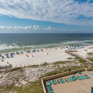 A panoramic view showcases the beach front with colorful umbrellas, sunbathers, and a sparkling pool area below