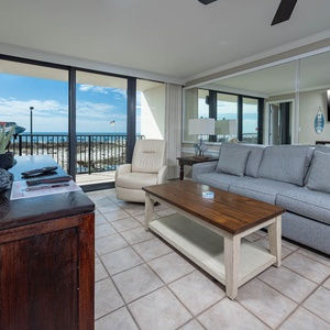The living area features a comfortable gray sofa, a sleek TV, and stunning beachfront views through large glass doors