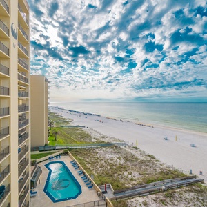 The view showcases a tranquil beachfront with white sand and the pool area, framed by a vibrant sky and nearby lounge chairs