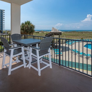 Balcony seating overlooks the pool area and lush greenery, with glimpses of the beach in the distance under a clear sky