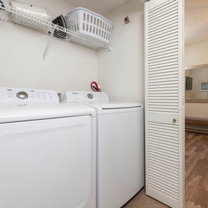 Washer and dryer set in a utility area with a wall-mounted shelf, leading to a cozy guest bedroom with a beach-themed print