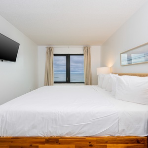 The master bedroom features a king-sized bed, a wall-mounted TV, and a stunning view of the beach through large windows