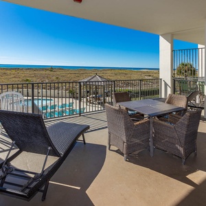 Balcony features outdoor seating with a dining table, overlooking a sparkling pool and the tranquil beach beyond