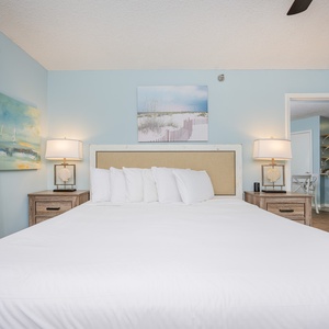 Bright and inviting master bedroom featuring a plush king bed, coastal artwork, and a glimpse of the modern kitchen beyond