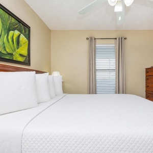This bedroom features a plush white bed, a wooden dresser, wall-mounted TV, tropical artwork, and natural light
