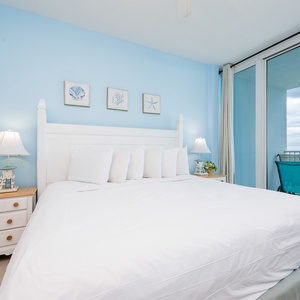 The bedroom features a serene blue wall, a cozy white bed, and a stunning beachfront view with direct balcony access