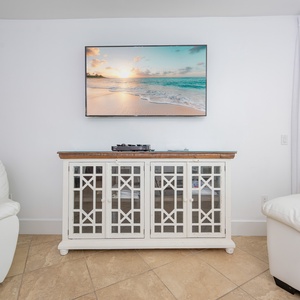 Living space featuring cozy white leather chairs, a stylish cabinet, and a large TV displaying a beach sunset