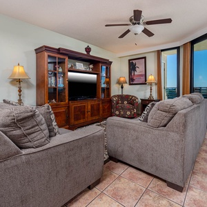 The living room features two cozy sofas, a wooden entertainment unit with a TV, and a stunning view through large glass doors